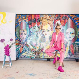 Del Kathryn Barton sits infront of a piece of her work in a pink suit with a vase of flowers to the left of her. 