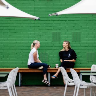 Two women sit on a bench seat at an outdoor cafe, having a conversation