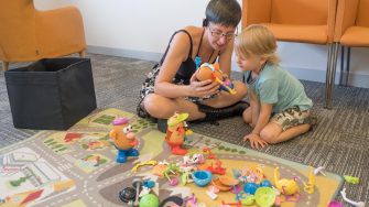 Child sitting with teacher on ground in front of playmat covered in toys