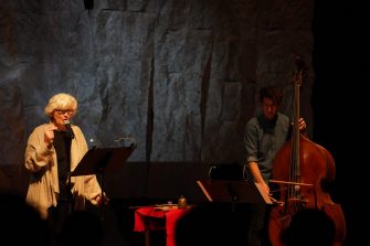 Two people, the woman on the left is speaking into a microphone while a man on the right is playing cello.