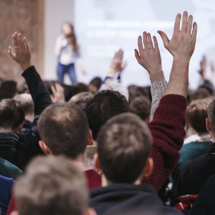 Adult students at large conference hall listen to woman speaker and getting hands up for answer on speaker's question