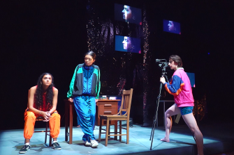 three students acting on stage, one holding a camera