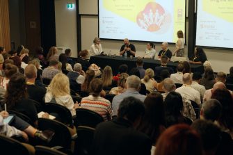 Six people are giving a presentation to many more people at a conference