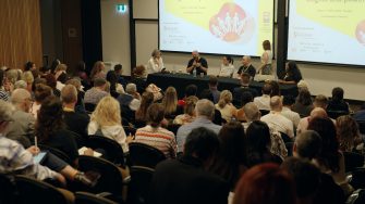 Six people are giving a presentation to many more people at a conference