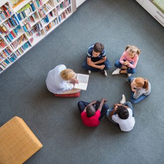 Teacher reading fairy tales to children sitting in a circle at library. Top view of librarian sitting with five multiethnic children on floor. Teacher reading book to girls and young boys at school.