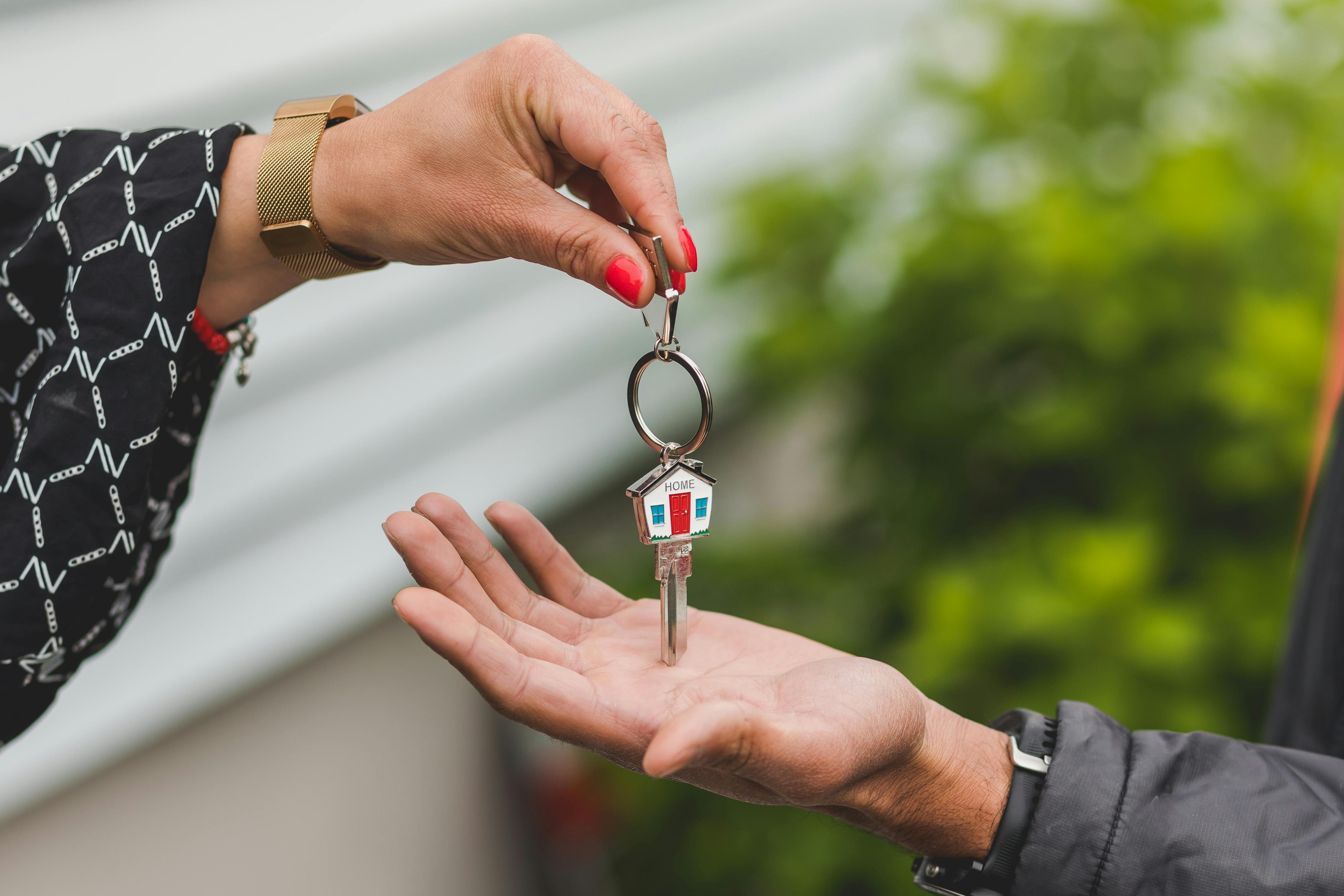 Close crop of an outstretched hand holding a house key pictured in front of a property.