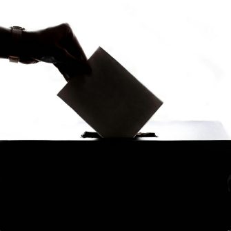 Close crop of a black and white silhouette of a hand casting a vote in a ballot box.