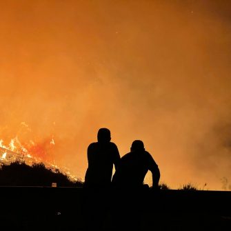Image depicts the silhouette of two crouching figures watching a nearby bushfire engulf the landscape, burning brightly against a flaming orange sky. 