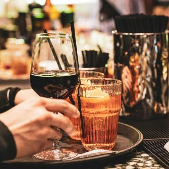A close crop image captures a pair of hands grasping a tray of alcoholic drinks at a bar.