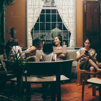 A large family sits in a living room talking to each other while a man in the background cooks dinner