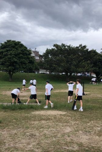 A group of school children accompanied by a teacher gather in a park under shade cloth to do sporting activity