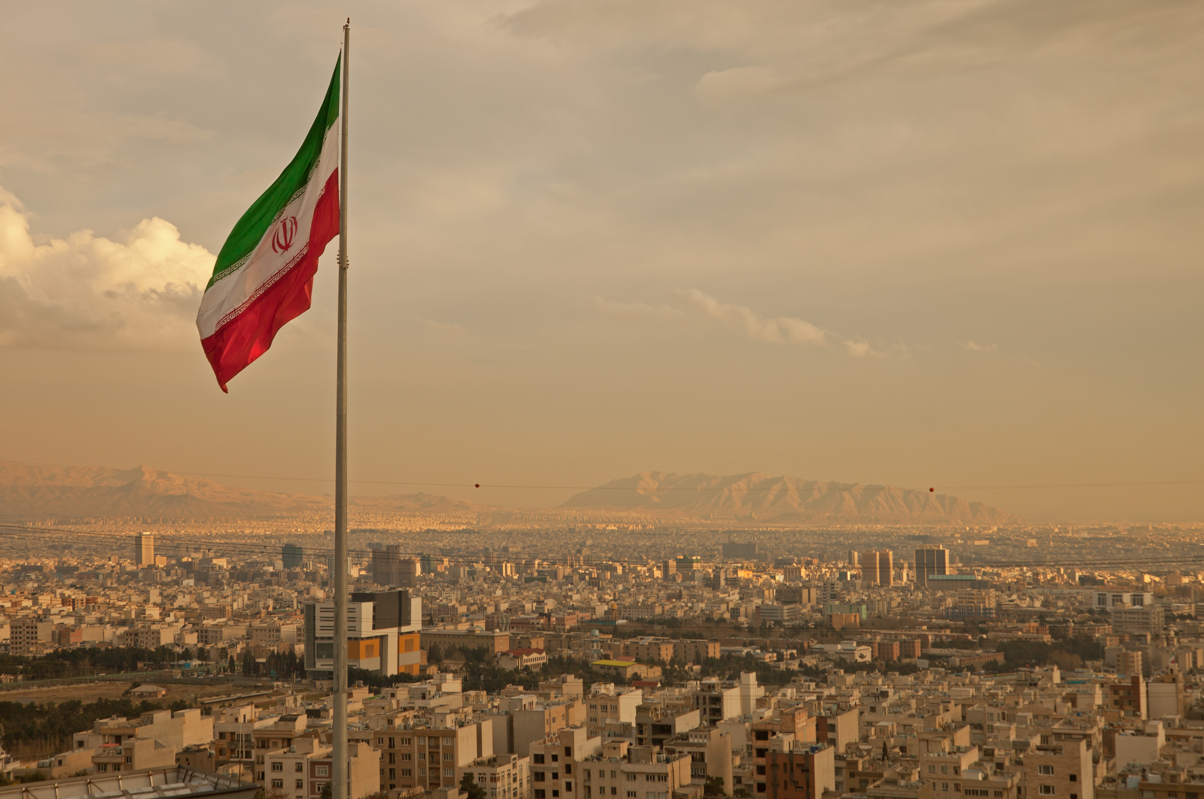 Iran flag waving  in the wind above skyline of Tehran lit by orange glow of sunset.
