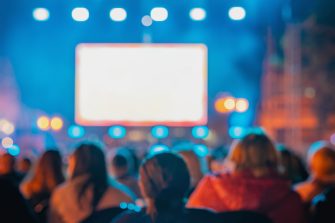 Rear view of blurred crowd watches large screen at night outdoor event..
