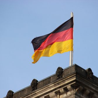 Close crop image of a German flag fluttering in the wind against a clear, blue sky, atop a historic building.