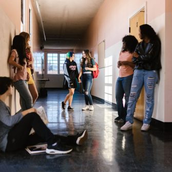 A wide camera angle captures a high school hallway populated by a loose group of students, some leaning against the walls, walking or a sitting position. 