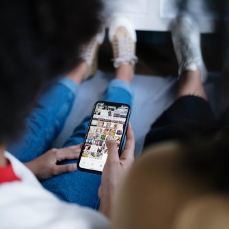 Two young people sit side by side facing away from the camera and gazing at a mobile phone device, with the focal point being the screen featuring a social media platform.
