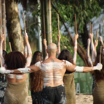 A group of Australian Aboriginal performers face away from the camera, captured in a dance in a verdant outdoor setting.
