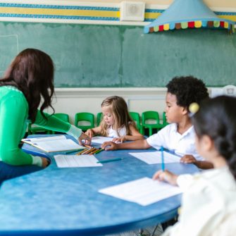 A teacher sits across from three of her students in a colourful classroom setting.