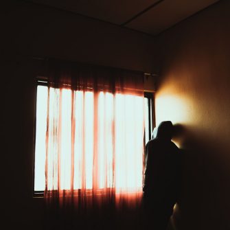 This stark contrast image depicts the silhouette of a hoodie-wearing figure standing inside a home dwelling, facing away from the camera and towards an illuminated window.