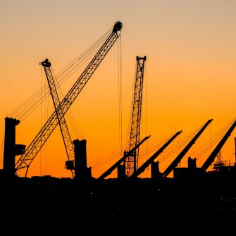 Several cranes silhouette an urban skyline against a vibrant orange sky.