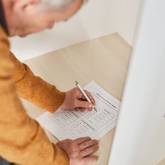 A high angle camera shot captures an older person leaning over a counter within a voting booth, hand poised over the voting ballot.
