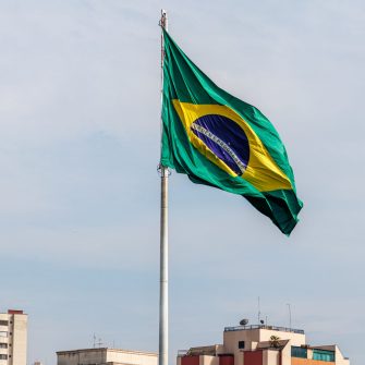 Brazilian flag flutters in the wind against a blue sky and cityscape skyline.