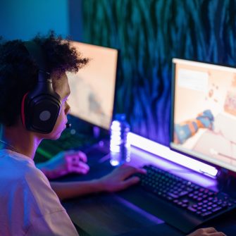 Two young boys sit at computer while gaming, wearing headsets and illuminated by their screens in a darkened room.