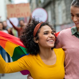 Young cheerful women on street enjoying holding gay pride flag during protest. Smiling multiethnic women enjoying victory after march on street for lgbt rights. Diversity, tolerance and gender identity concept.