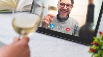 A hand holds a glass of white wine toasting a man on an iPad screen holding a glass of white wine 