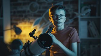 Smart nerd guy with glasses watching stars with a telescope in his bedroom at night, science and childhood concept