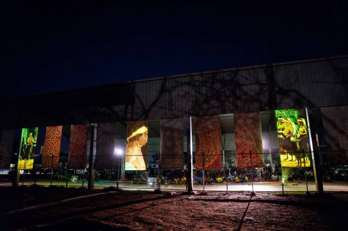 A dark building with illuminated banners in front. A group of people in brightly coloured shirts sit on the ground behind the banners.