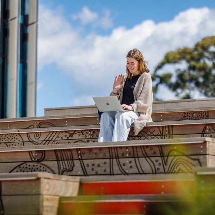 Student waving at laptop while sitting on stairs.