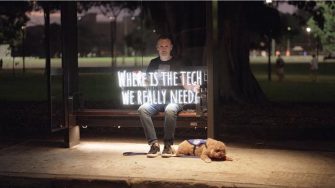A man wearing jeans and a t-shirt sits in the glow of a bus stop at night, holding a neon sign that reads “where is the tech we really need”, with a fluffy light brown assistance dog lying by his feet