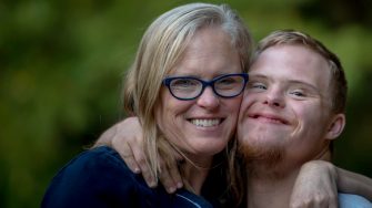Two people share a close, smiling embrace in an outdoor setting with a blurred green background.