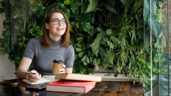 Female Creative smiling and contemplating idea holding coffee cup in modern office shot through window and green plants