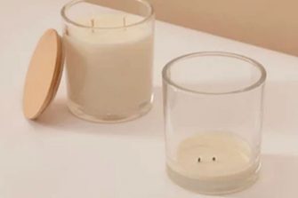 Two glass candle jars with white wax and twin wicks sit on a table, with a wooden lid beside one jar.