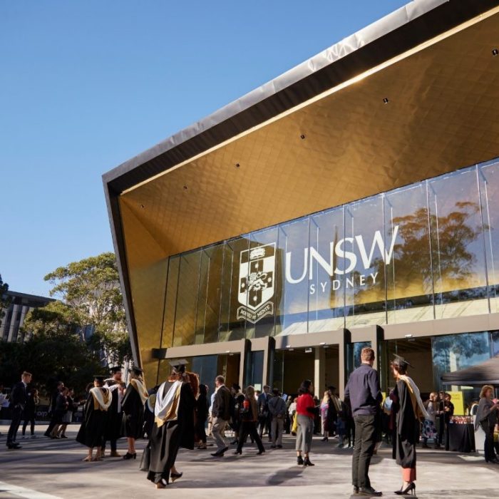 Graduates in academic gowns and visitors gather outside the golden UNSW Sydney building entrance displaying the UNSW Sydney logo and crest on a clear day.
