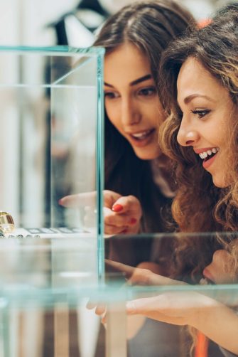 Two women smile as they admire jewelry in a glass display case, one pointing excitedly at the shiny rings inside.