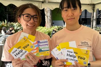 Two women are smiling at the camera, holding Health and Wellbeing pamphlets. One of them is wearing a Health and Wellbeing Ambassador t-shirt.