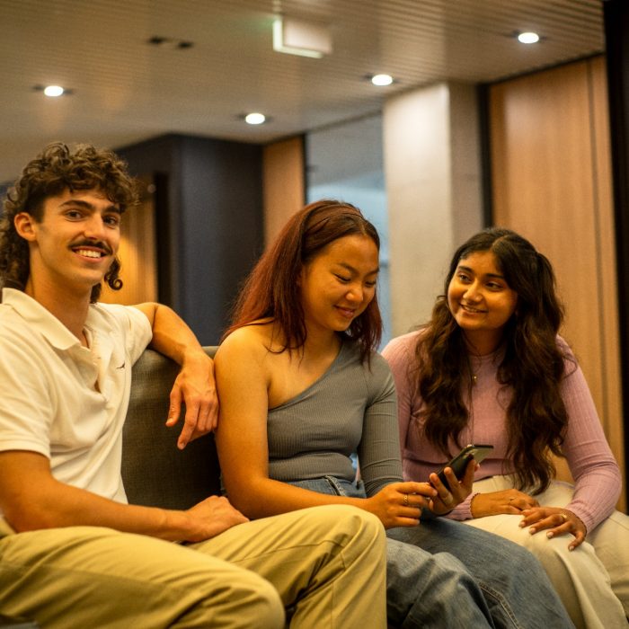 Three students smiling and looking at one of their phones while sitting on a bench indoors on campus