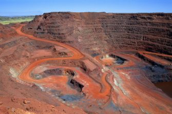 "Looking down on a large open cut iron ore mine in Australiaa's remote Pilbara region.  At the bottom of the pit at the end of the red winding road, a dump truck is being loaded with ore.  Other mine vehicles appear tiny in the huge pit.  Beyond the open cut are green hills.  Taken with permission during a guided tour of the mine."