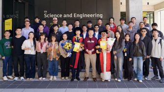 A large group of UNSW students and staff pose outside the Science and Engineering building, with two graduates in academic gowns holding flowers and certificates.