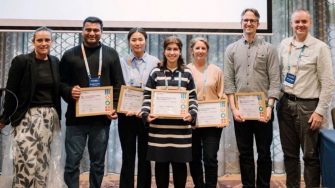 Seven people stand together at an awards ceremony, with four holding framed certificates and smiling for the camera.