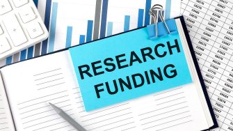 'Research funding' on a post-it note stuck to a notebook with charts and graphs in the background.