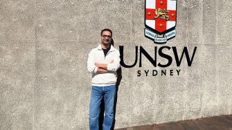 A man stands with his arms crossed beside the UNSW Sydney crest and signage at the Kensington campus.