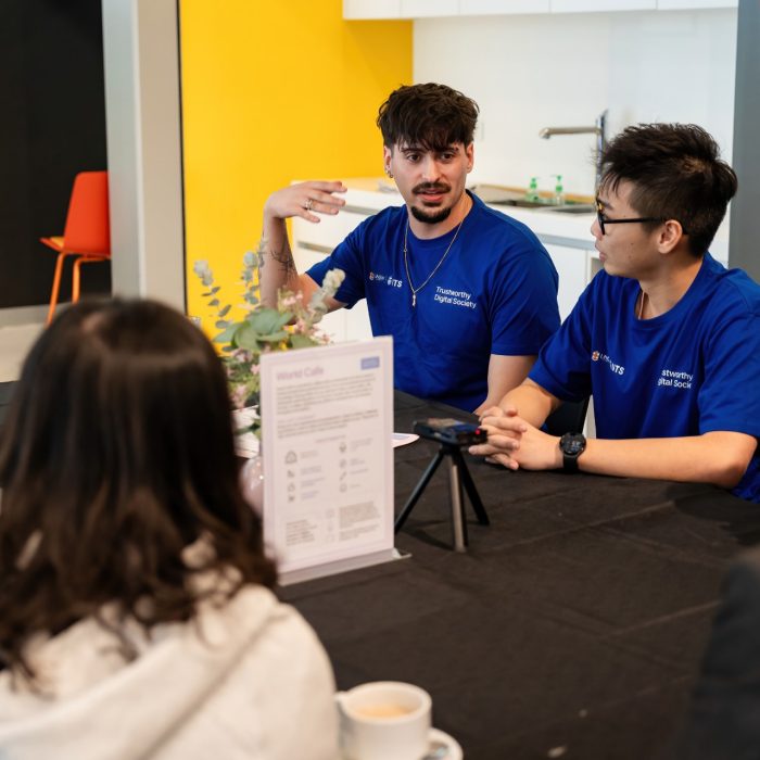 A participant in a blue T-shirt gesturing while speaking to others around a table at the UNSW–UTS TDS Student Forum.
