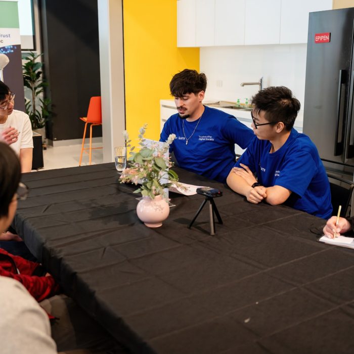 Two participants wearing blue T-shirts talking with others around a table at the UNSW–UTS TDS Student Forum.