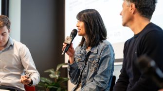 A panellist in a denim jacket speaking into a handheld microphone while seated beside other speakers at the UNSW–UTS TDS Student Forum.