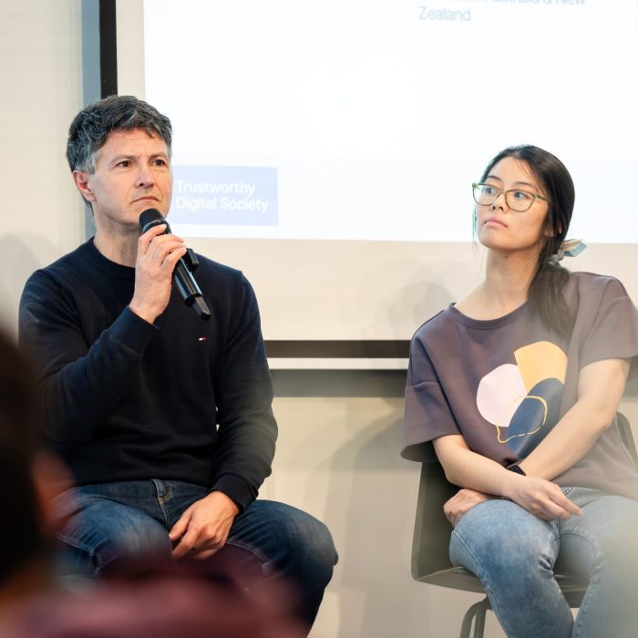 A panellist in a navy jumper speaking into a microphone beside another seated speaker at the UNSW–UTS TDS Student Forum.