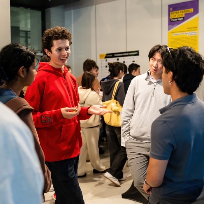 An attendee in a red hoodie gesturing while talking with a standing group at the UNSW–UTS TDS Student Forum.